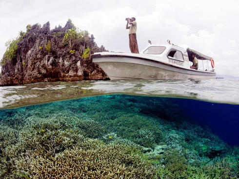 A person stands with binoculars on the front of a boat amid a coral reef.