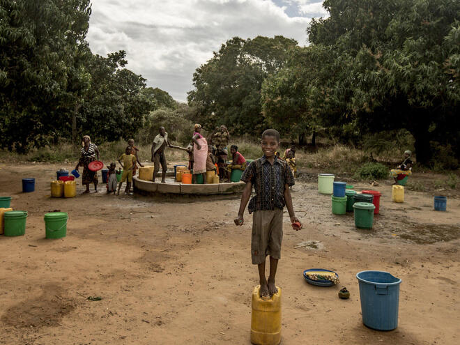 Boy stands on a container used for carrying water, with women gathering water in the background.