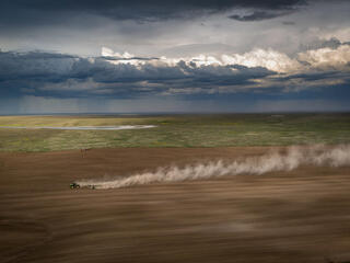 A tractor plows a vast field with a rain storm in the distant background