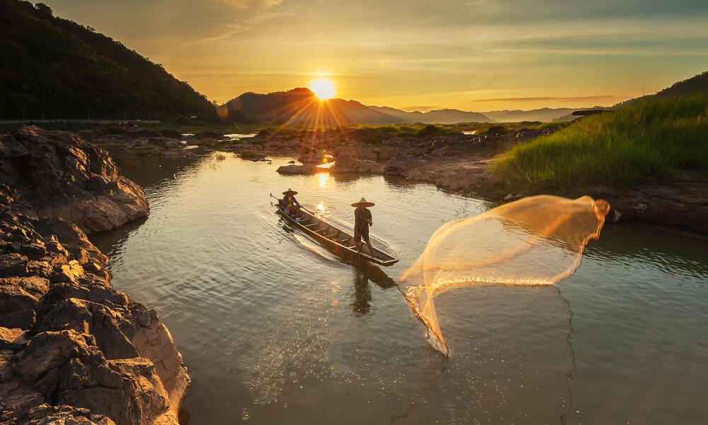 Two people cast a fishing net into the Mekong River as the sun rises