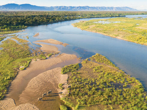 Wide angle aerial photo showing elephants crossing river in foreground and mountains in background