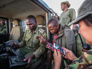 Group of people stand around an open jeep looking at a hand-held device with keyboard